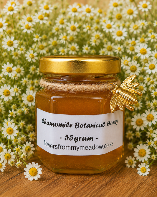 A 55g glass jar of chamomile botanical honey with a gold lid and a white label, decorated with a gold bee ornament and wrapped in twine, set against a background of chamomile flowers on a wooden surface.