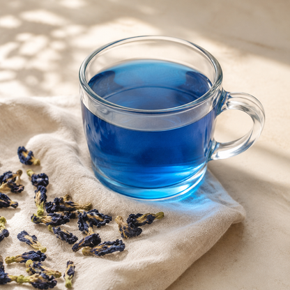 Clear glass mug containing vivid blue Butterfly Pea Flower tea beside dried flowers on warm ivory linen.