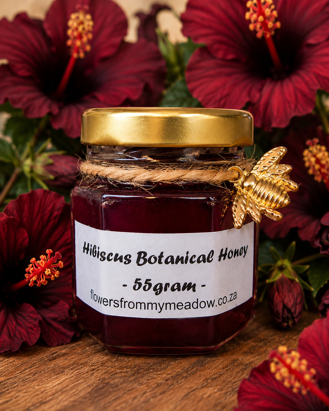 A 55g glass jar of hibiscus botanical honey with a gold lid and a white label, decorated with a gold bee ornament and wrapped in twine, set against a background of hibiscus flowers on a wooden surface.