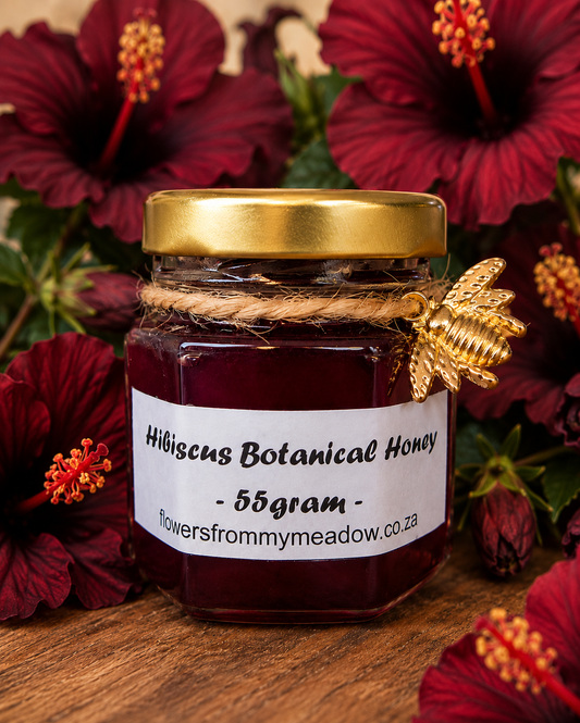 A 55g glass jar of hibiscus botanical honey with a gold lid and a white label, decorated with a gold bee ornament and wrapped in twine, set against a background of hibiscus flowers on a wooden surface.