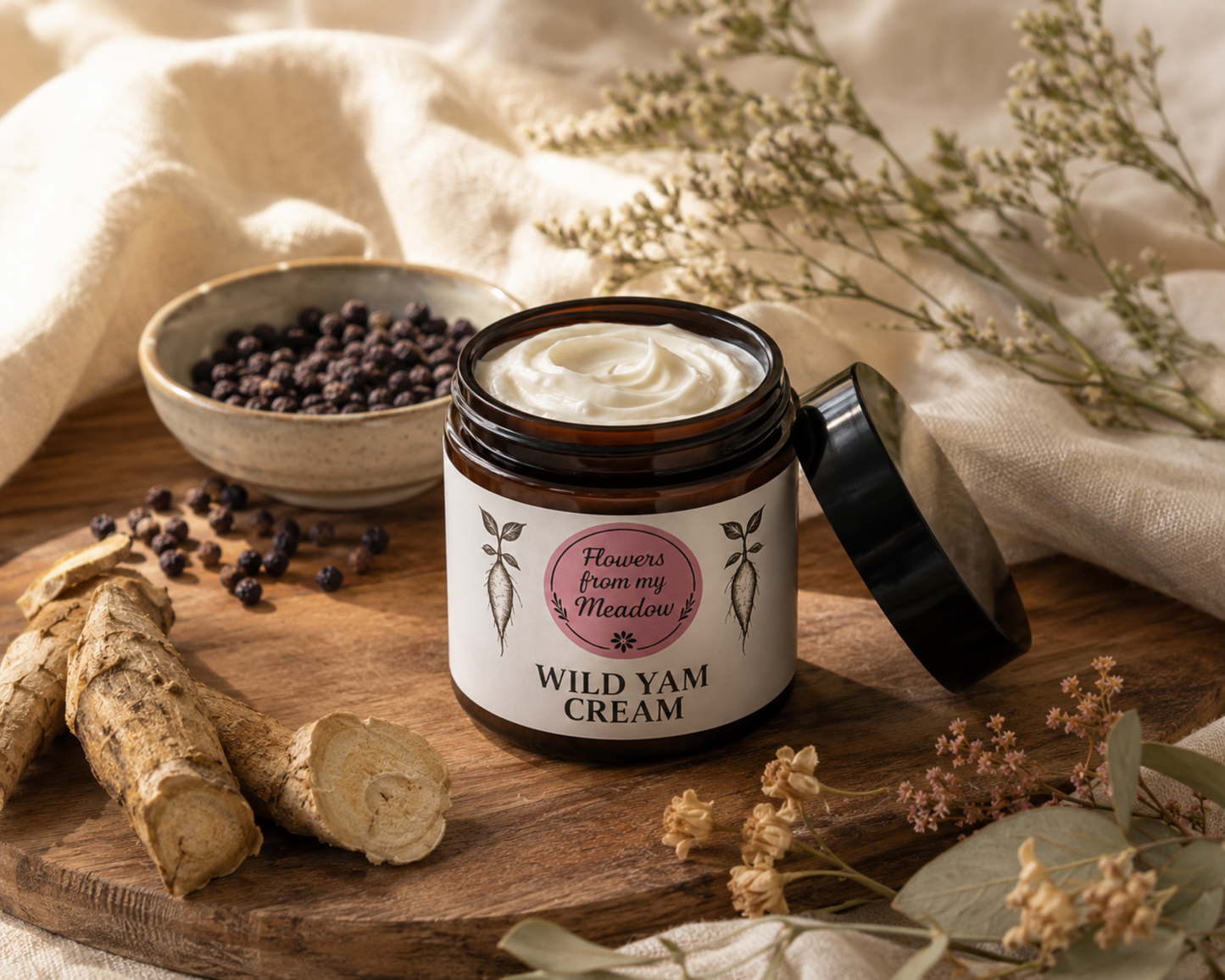 An open jar of Flowers from my Meadow Wild Yam Cream sits on a wooden board with raw yam roots, dried flowers, and a small bowl of dark yams, set against a soft beige fabric backdrop.
