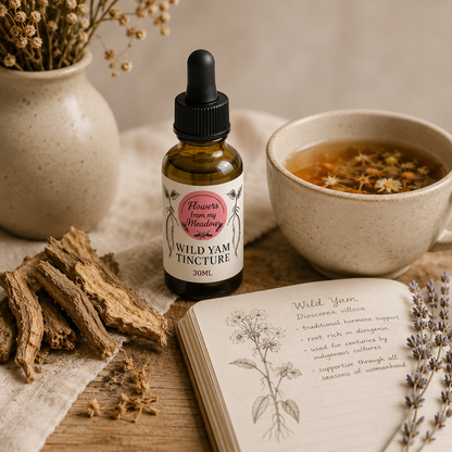 A small amber dropper bottle labeled 'Flowers from My Mindsters Wild Yam Tincture 30ml' sits on a wooden surface with dried flowers, a cup of herbal tea, bark pieces, and an open notebook with botanical notes.