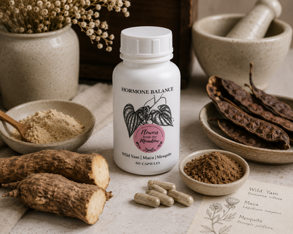 A white bottle labeled 'HORMONE BALANCE' with botanical artwork and a pink flower emblem, styled on a tabletop with wild yam roots, maca powder, capsules, dried pods, bowls, and a mortar and pestle.