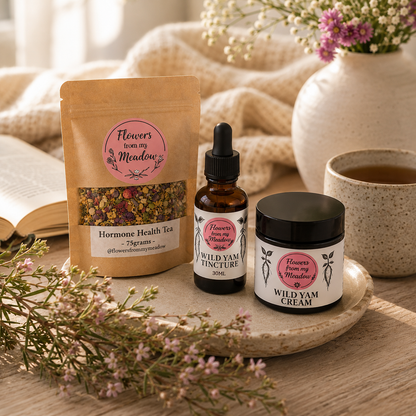Three herbal wellness products from Flowers from my Meadow are arranged on a wooden tray with a ceramic mug, flowers, and a book in a warm, cozy setting. The products include a brown kraft stand-up pouch of Hormone Health Tea, an amber glass dropper bottle of Wild Yam Tincture, and a black-lidded jar of Wild Yam Cream.