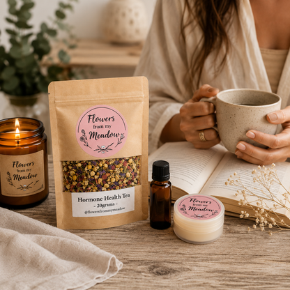 A styled tabletop scene showing a person holding a ceramic mug beside a stand-up pouch of 'Flowers from my Meadow' hormone health tea, a small amber bottle, and a round tin of the same product. The setting includes an open book, candle, and dried flowers on a wooden surface.