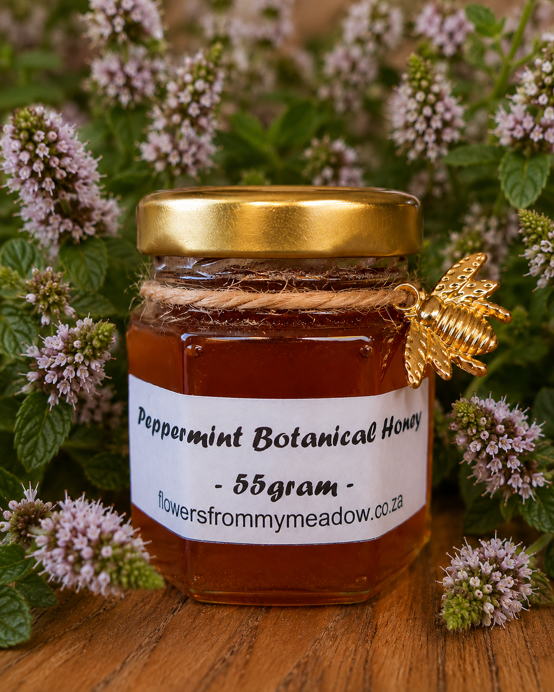 A 55g glass jar of peppermint botanical honey with a gold lid and a white label, decorated with a gold bee ornament and wrapped in twine, set against a background of flowering peppermint on a wooden surface.