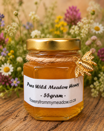 A 55g jar of Pure Wild Meadow Honey on a rustic wooden surface, with a white label and a decorative gold bee ornament tied with twine. A blurred background of wildflowers and greenery creates a natural setting.