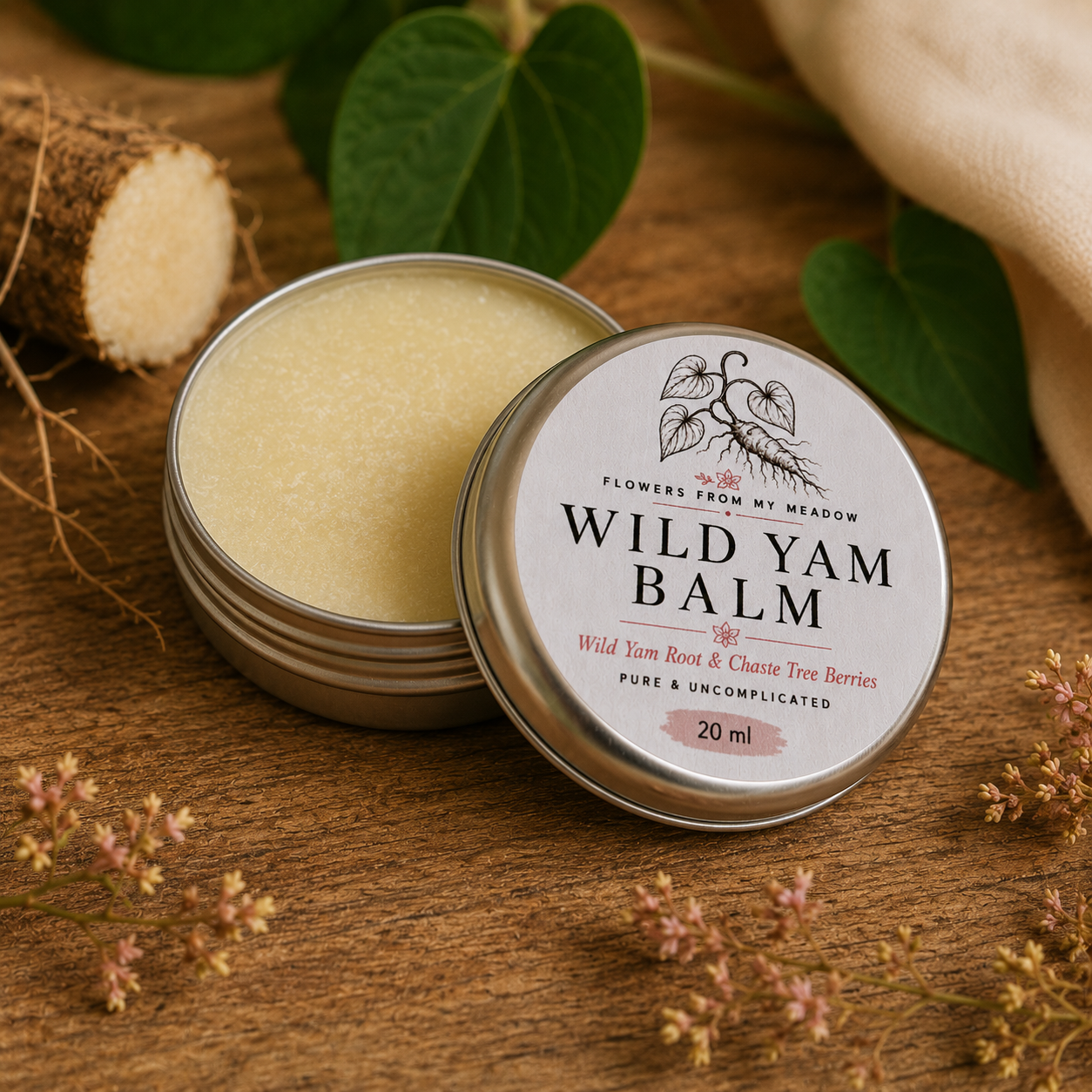 An open round metal tin of Flowers from my Meadow Wild Yam Balm sits on a wooden surface next to its labeled lid. The balm is a pale creamy yellow, with green leaves, dried flowers, and a piece of root visible around it, creating a natural, rustic setting.
