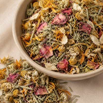 A close-up of a ceramic bowl filled with dried flower and herb tea blend, including pink petals, yellow-orange blossoms, and greenish stems, set on a beige fabric surface.